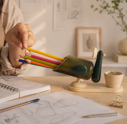 Person holding colored pencils over a green pencil holder on a desk with drawing materials.